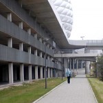 Person, die an einem modernen Gebäude mit der markanten Allianz Arena im Hintergrund vorbeigeht. Fritten, Fussball & Bier - www.soccer-warriors.de