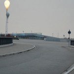 Ein bewölkter Tag an einer leeren, kurvigen Straße, die zur Allianz Arena führt, mit Straßenlaternen und einer Schranke, möglicherweise auf einer Brücke oder Überführung. Fritten, Fussball & Bier - www.soccer-warriors.de