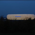 Beleuchtete Allianz Arena in der Abenddämmerung mit umliegender Landschaft. Fritten, Fussball & Bier - www.soccer-warriors.de