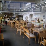 Eine ruhige, moderne Cafeteria mit Holztischen, einer Essenstheke im Hintergrund und reflektierenden Fenstern, die teilweise das gespenstische Spiegelbild einer Person im Grünwalder Stadion zeigen. Fritten, Fussball & Bier - www.soccer-warriors.de