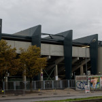 Modernes Gebäude mit freiliegenden Strukturelementen und einer Straßenbahnlinie im Vordergrund, teilweise verdeckt durch einen Metallzaun und eine Werbetafel in der Nähe des Grünwalder Stadions. Fritten, Fussball & Bier - www.soccer-warriors.de